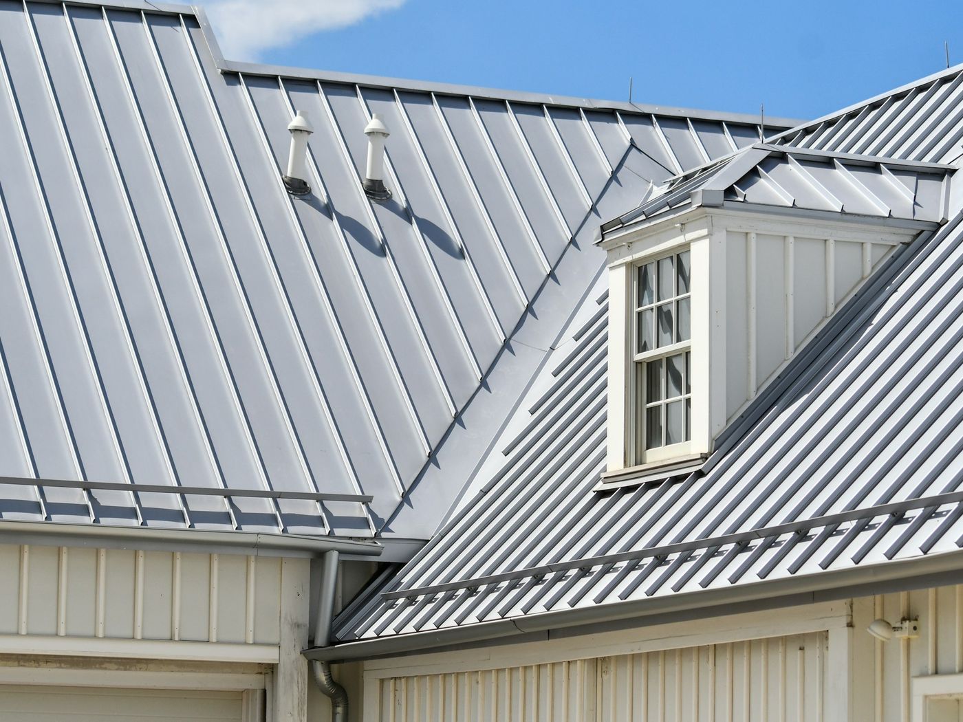 Two grey standing-seam metal roof planes meeting at a valley, with a white dormer and lap-sided wall