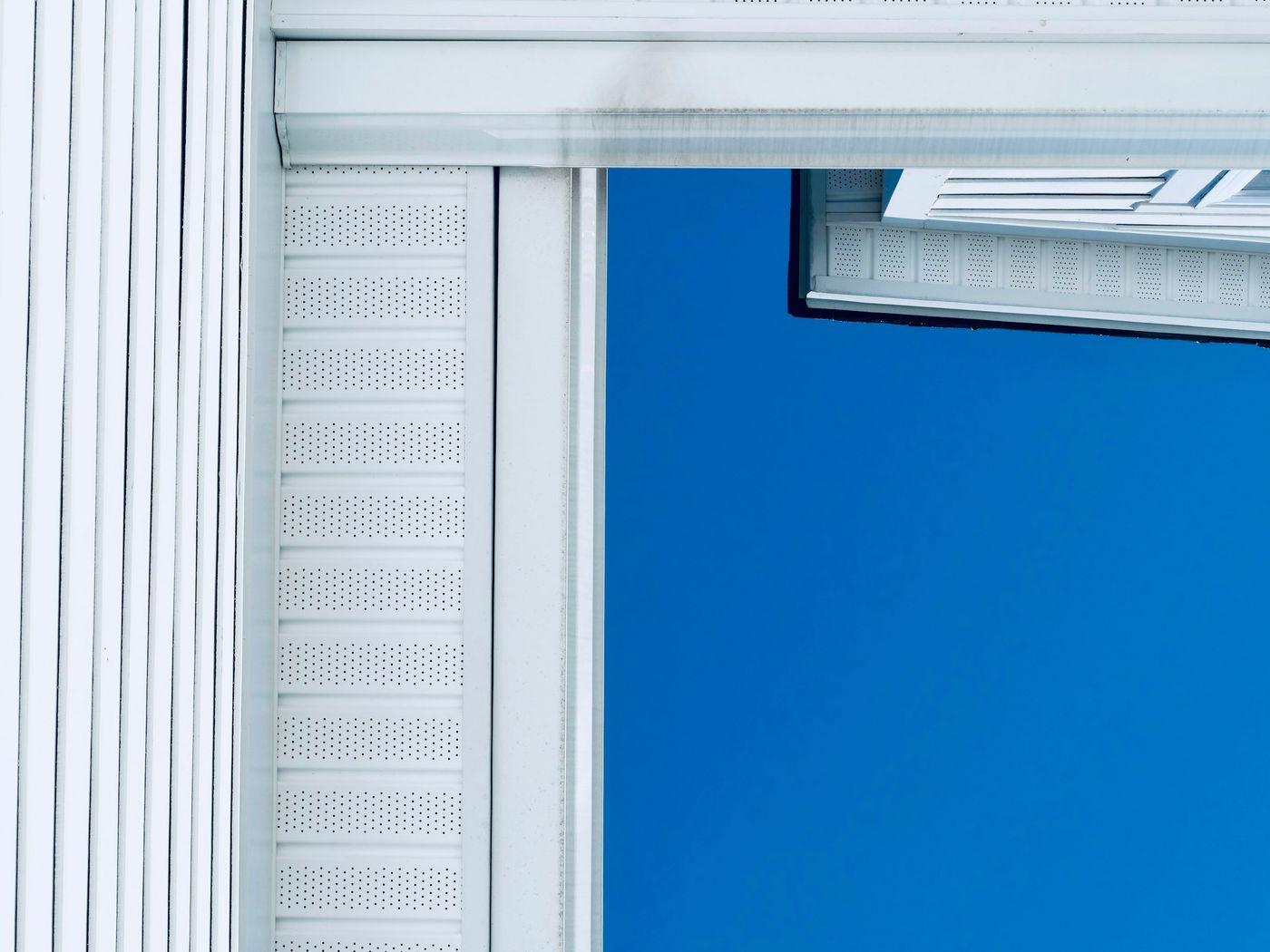 Close-up of a white perforated vented soffit panel and fascia beneath a roof edge, against a deep blue sky